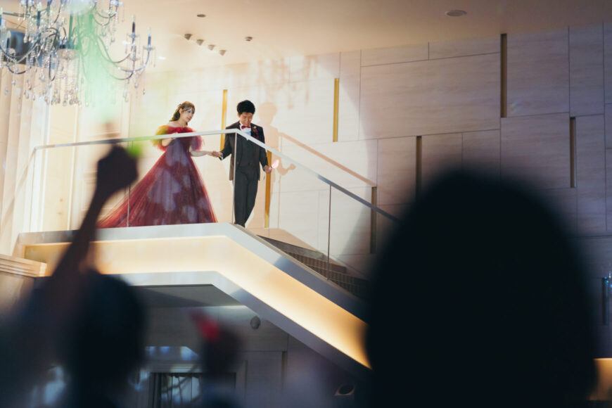 大階段から再入場する赤いカラードレスの新婦と新郎